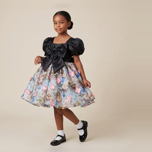 Girl wearing a black satin girls party dress with puff sleeves, an oversized front bow and a blue 3D floral skirt.