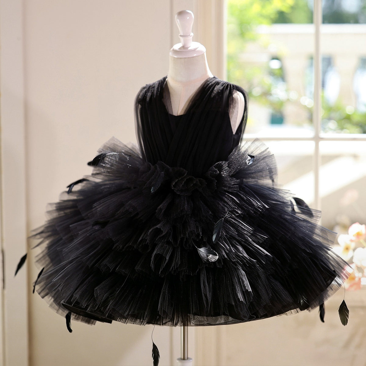 Black dress for girls in pleated tulle with a flower and feather waist detail, angled front view, for parties and pageants.