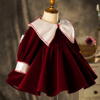Burgundy velvet dress with pom-pom trimmed collar and cuffs, angled view, for a winter celebration.