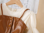Close-up of the brown faux leather vest with thin straps and snap buttons over the cream chiffon girls dress, designed for parties.