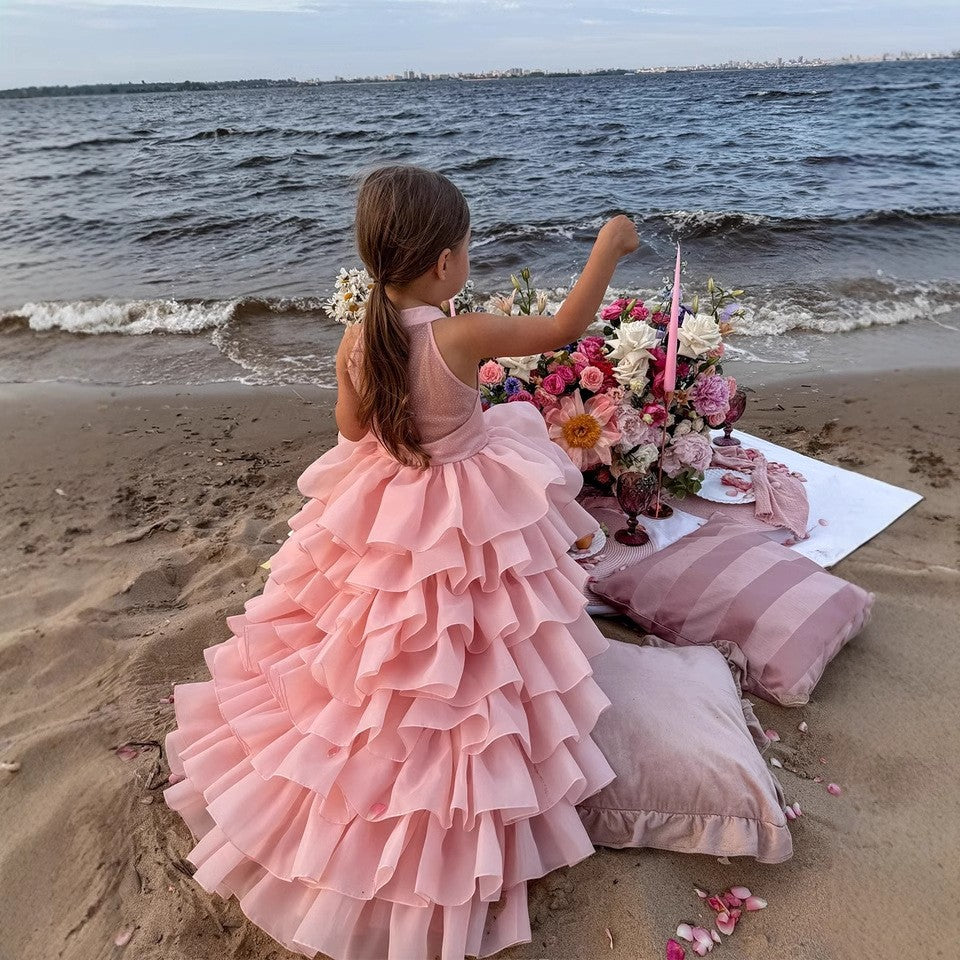 Blush pink princess dress for girls with stacked organza ruffles, seated beside a flower table on the sand.