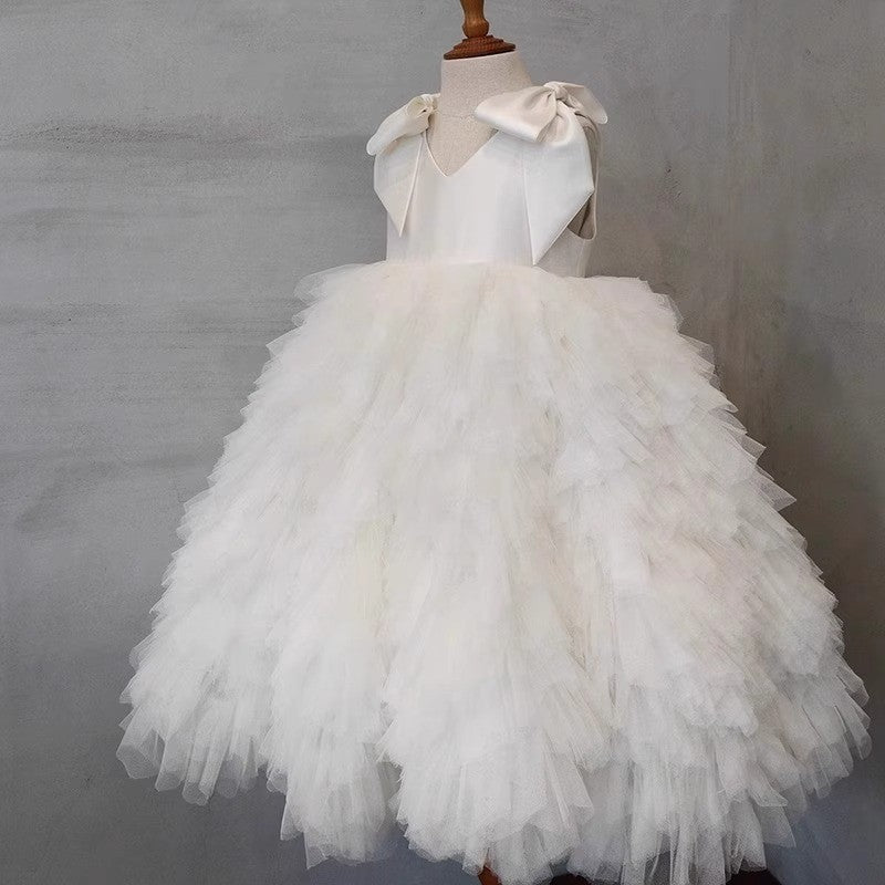 White dress for girls in tiered tulle with satin shoulder bows, styled against a soft grey backdrop for ceremony photos.